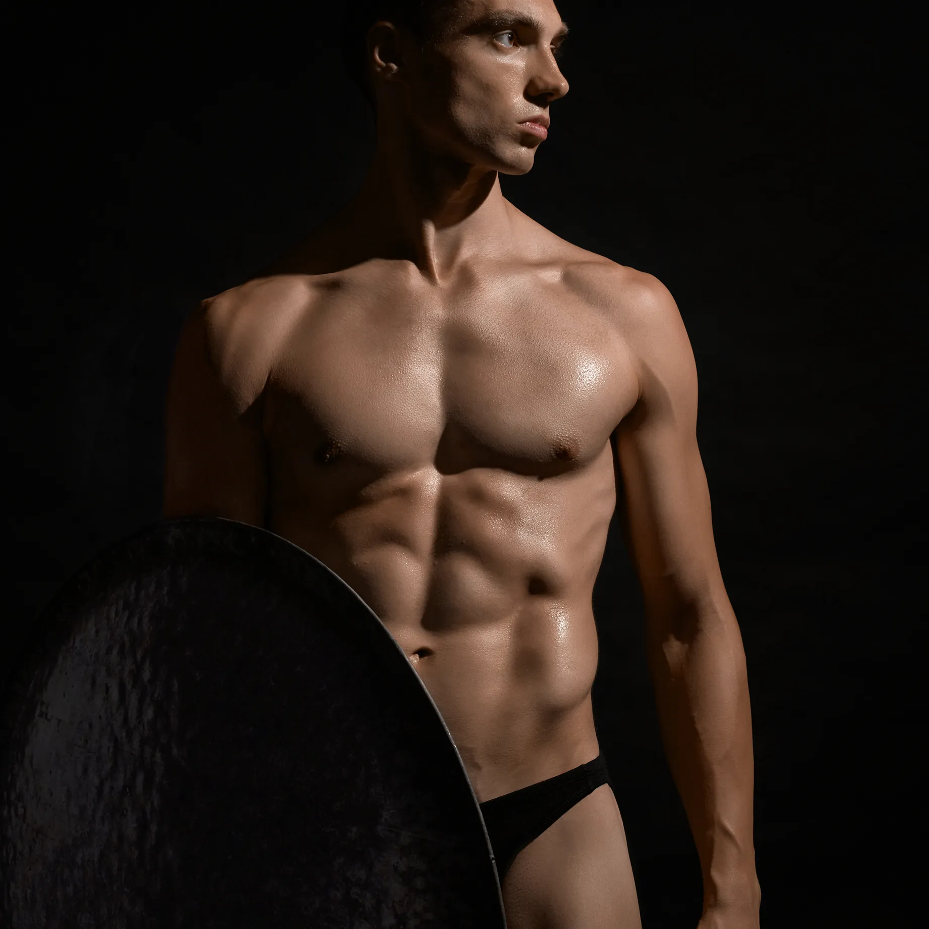 Homme musclé torse nu, éclairage sombre, tenant un grand bouclier en métal, regardant vers la droite, portant un slip noir